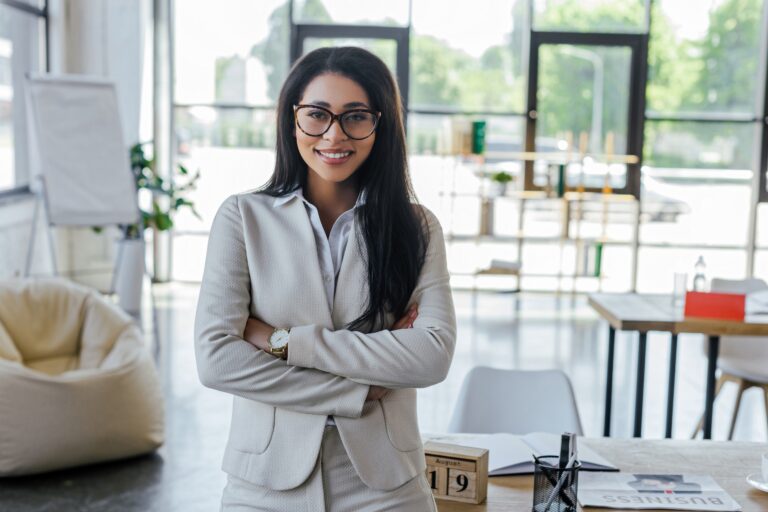 happy businesswoman in glasses standing with crossed arms near table in office